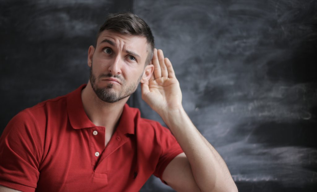Man in red shirt in front of chalkboard with hand cupped to his ear in a listening position