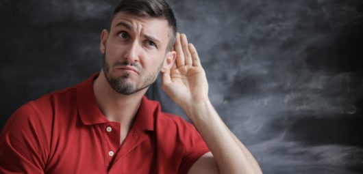 Man in red shirt in front of chalkboard with hand cupped to his ear in a listening position