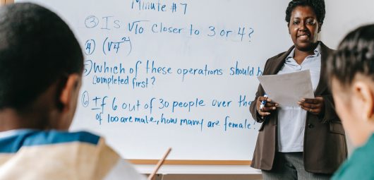 Math teacher standing in front of room with problems written on white board and students are at their desks working