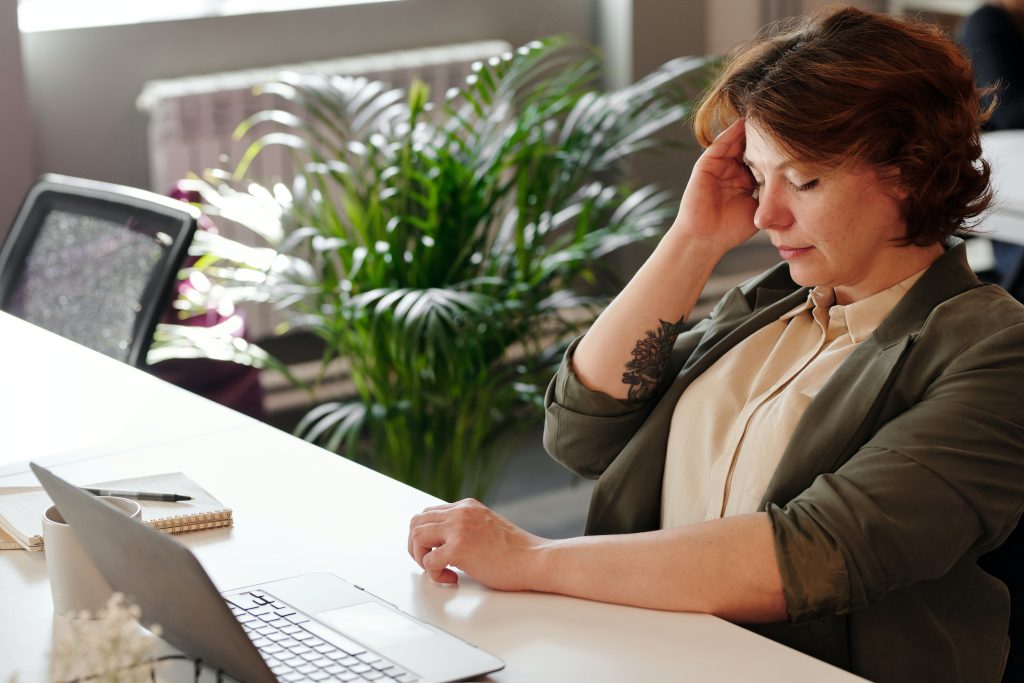 Photo of woman sitting in front of her computer, with a hand on her head, looking tired and overwhelmed