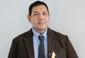 man standing in front of camera, slight smile, holding documents in his hands