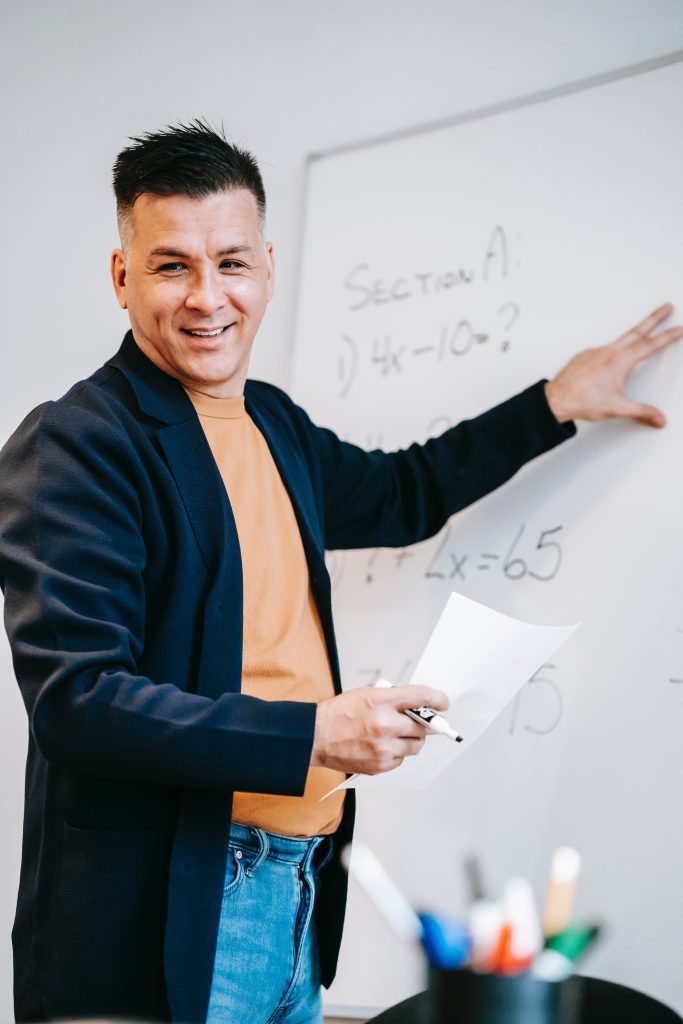 Photo of smiling man pointing to white board while holding a piece of paper and marker