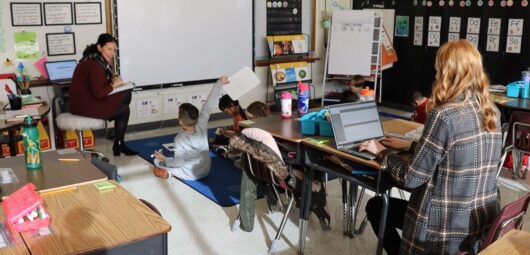 A teacher working with kids in her classroom while another teacher observes and takes notes on a computer in the NEE POWERHub peer observation tool