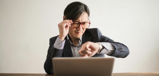 Man sitting in front of his computer, checking his watch while holding his eyeglasses